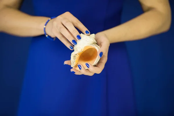 ブルーのドレスの前に小さな海カタツムリの殻を持って完璧な青いマニキュアで美しい女性の手をバック グラウンドとして使用できます — ストック写真