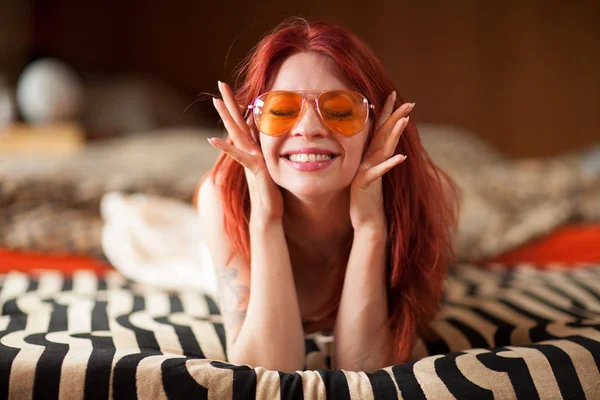 pretty young woman with red hair and sunglasses lies relaxed and laughing on the couch, emotion concept shoot