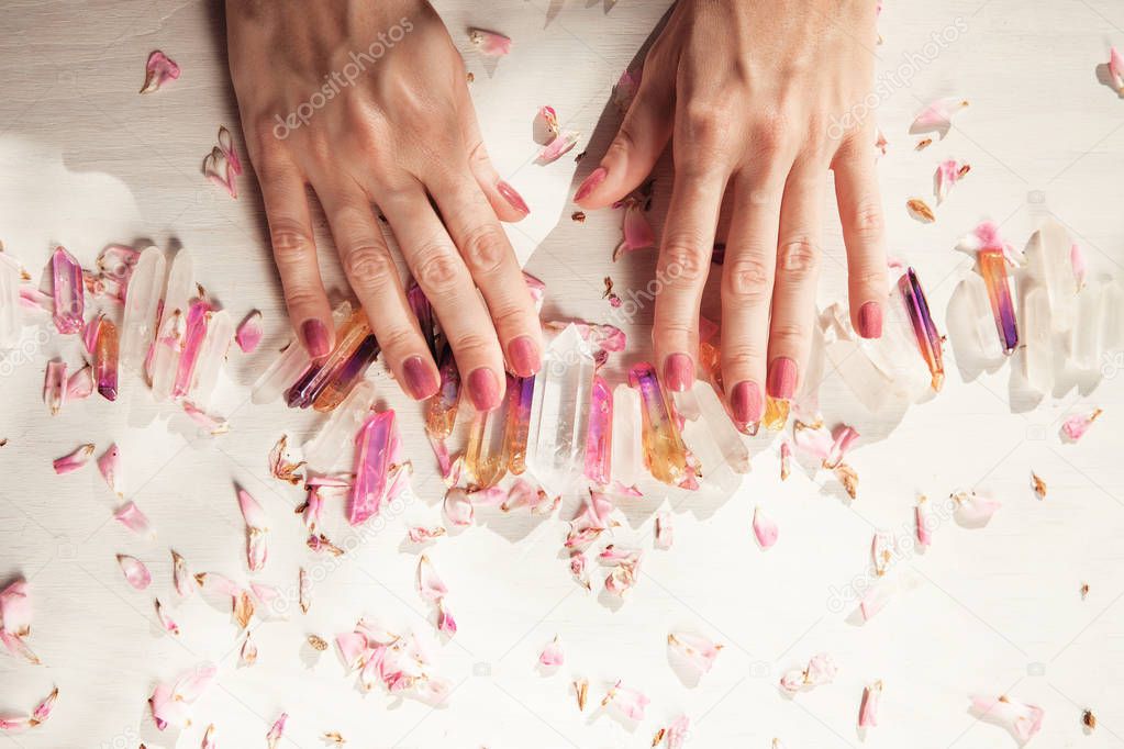 Beautiful woman hands with perfect violet nail polish on white wooden background holding little quartz crystals, decorated with rose leaves, healing crystal concept shoot, can be used as background