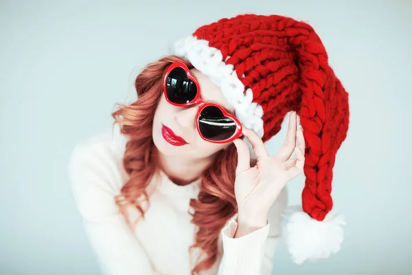 サンタ リトル ヘルパー サンタ クロースの帽子 赤い口紅とハート形のサングラスの完璧なメイクアップ 愚かな 面白い気分に美しい幸せな若い女 — ストック写真