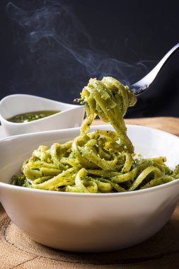 Pesto soslu makarna tabağı dumandan çıkıyor. Peynir, pesto ve fesleğen yapraklarıyla çevrili kaseler.