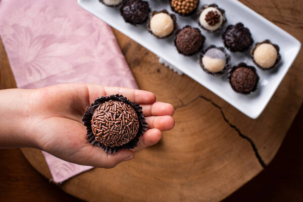 Рука ребенка держит Бригадейро. Типичный бразильский сладкий. Многие типы бригадиров вместе на заднем плане