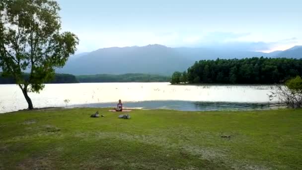 Girl sits on mat meditates near tree at lake — Stock Video