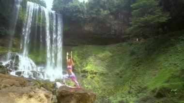 Ince sarışın kız yoga poz rock againat şelale güneşli nehir kıyısında üzerinde değiştirmek için el yükseltir