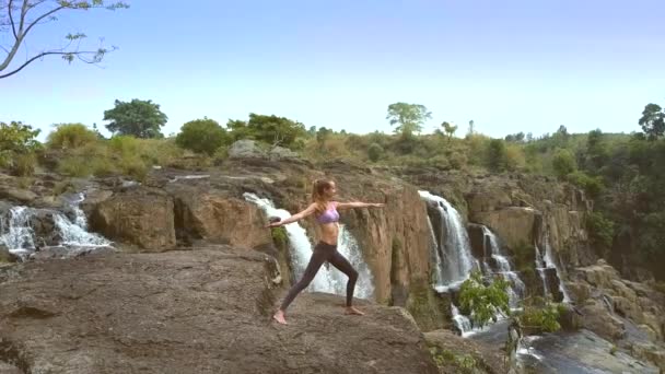 Blondine steht in Yoga-Pose auf flachem Felsen am Wasserfall — Stockvideo