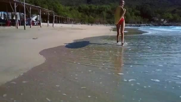 Chica camina a lo largo de la costa atada a la tabla de surf contra las plantas — Vídeos de Stock