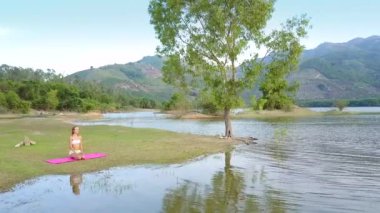 resimsel yuvarlak görünümü güzel Bayan pembe mindere oturur ve göl su hills karşı büyük kıvırcık ağacı yansıtır
