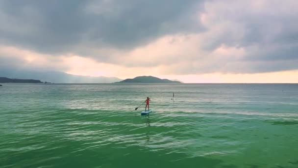 图片上部运动对小女孩站立在 Paddleboard 在蔚蓝的海洋之间对海岛和多云天空 — 图库视频影像