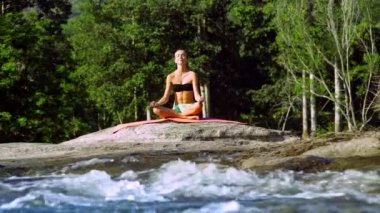 Genç kadın halıda oturuyor ve dere kıyısında meditasyon yapıyor. 