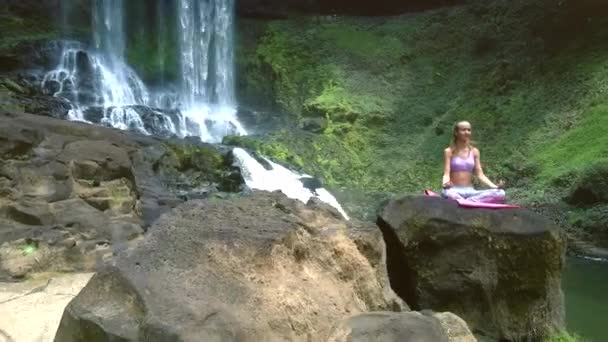 Blond meisje zit in lotus pose op rots door waterval in het park — Stockvideo