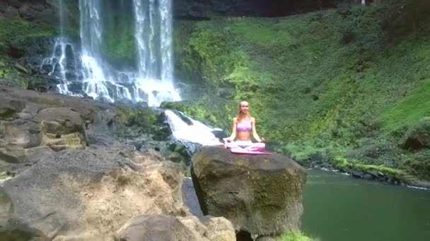 Drone elimina de chica sentada en pose de loto en piedra por río y cascada — Vídeos de Stock