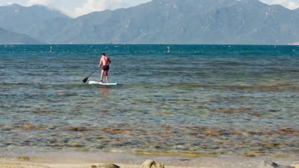 Хлопець весла на дошці на океанічному пляжі проти пагорбів — стокове відео