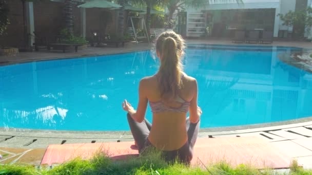 Menina senta-se em ioga pose lótus por piscina vista traseira — Vídeo de Stock