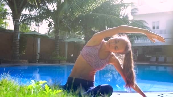Jovem mulher senta-se em ioga pose dobra as mãos para esquerda pela piscina — Vídeo de Stock