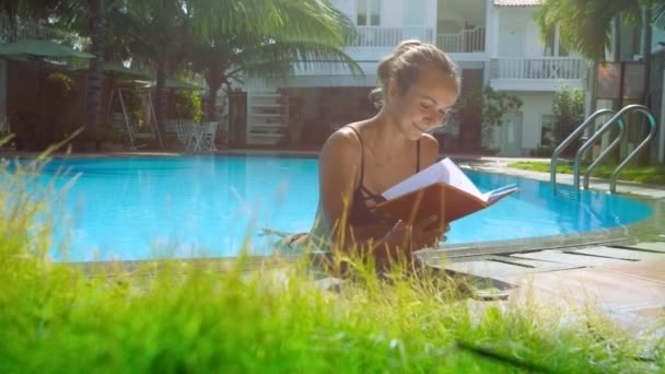 Menina loira de biquíni lê livro na piscina apoiando-se na barreira — Vídeo de Stock