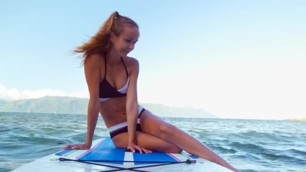 Blond girl puts feet into water from paddle board in ocean — Stock Video