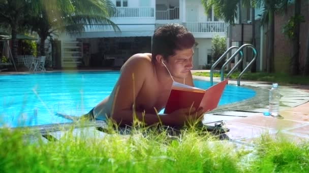 Молодий чоловік читає паперовий щоденник слухати музику по краю басейну — стокове відео