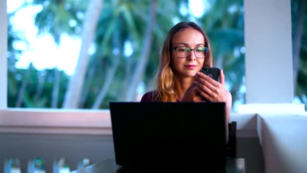 Girl in glasses surfs internet on phone against palm trees — Stock Video
