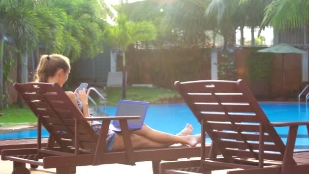 Жінка зі смартфоном і ноутбуком сидить у кріслі біля басейну — стокове відео