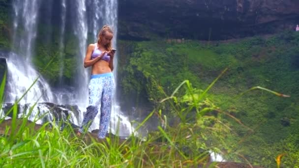 Menina surfa internet no telefone atrás da grama contra a cachoeira — Vídeo de Stock