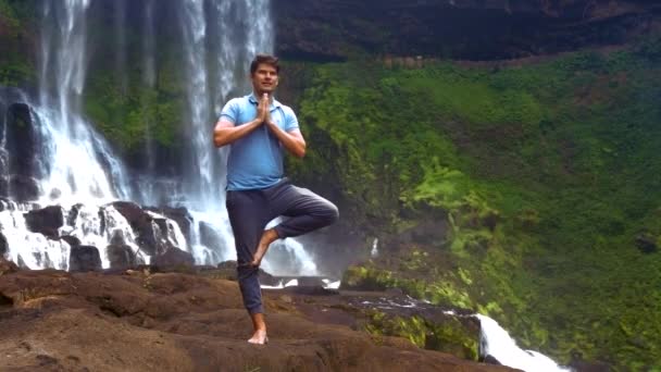 Mann hält Pose auf Beinfalten Handflächen auf der Brust am Wasserfall — Stockvideo