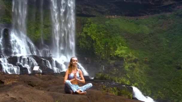 Блондинка сидит в позе йоги Пранаямы на скале у водопада — стоковое видео
