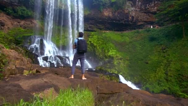 Chico de la parte trasera con la mochila se para en la cascada relojes de roca — Vídeos de Stock