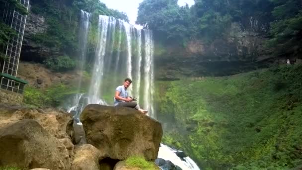 Cara senta-se na rocha com flycam controle remoto sorrindo por cachoeira — Vídeo de Stock