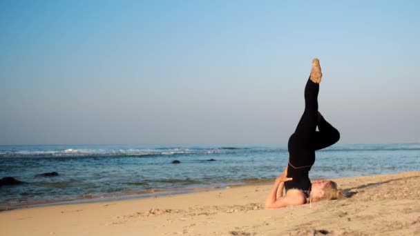 Стройная девушка в спортивном костюме медитирует на пляже под голубым небом — стоковое видео