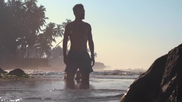 Hombre guapo en pantalones cortos silueta soportes en aguas poco profundas — Vídeos de Stock