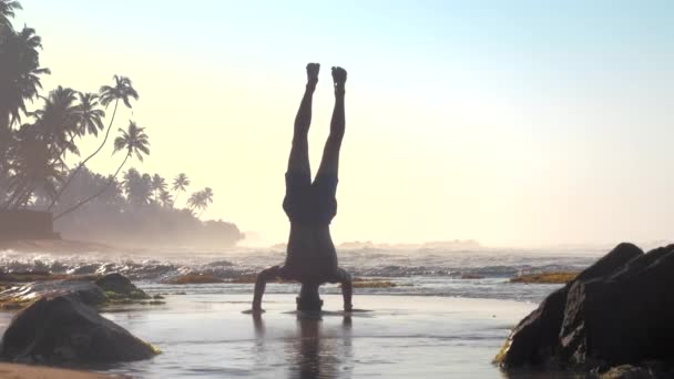 Αθλητής στέκεται στα χέρια και το κεφάλι κατά του αφρού κύματα του ωκεανού — Αρχείο Βίντεο