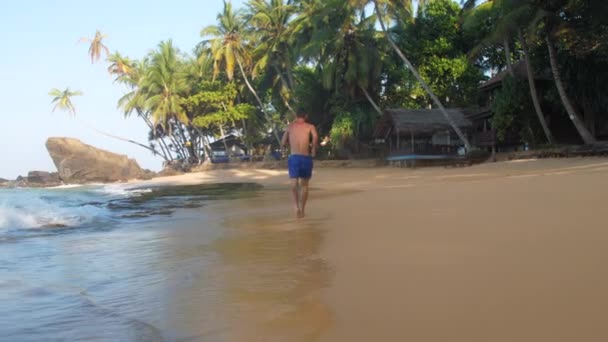 Desportivo muscular homem em calções azuis corre ao longo da costa — Vídeo de Stock