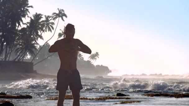 Hombre guapo medita en la costa del océano contra las olas espumosas — Vídeos de Stock