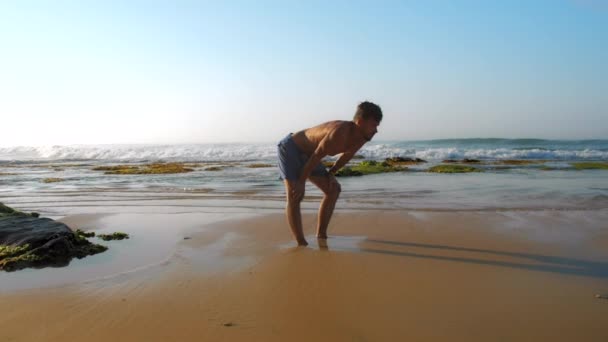 Homem forte em shorts estende as pernas em pé na areia molhada — Vídeo de Stock