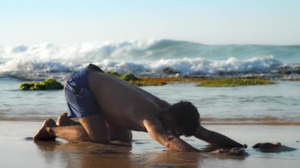 Muscular gymnast in blue shorts practices yoga on beach — Stock Video
