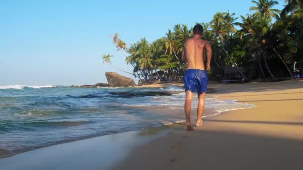 Muskularny gimnastyk biegnie wzdłuż piaszczystej plaży oceanu powolny — Wideo stockowe