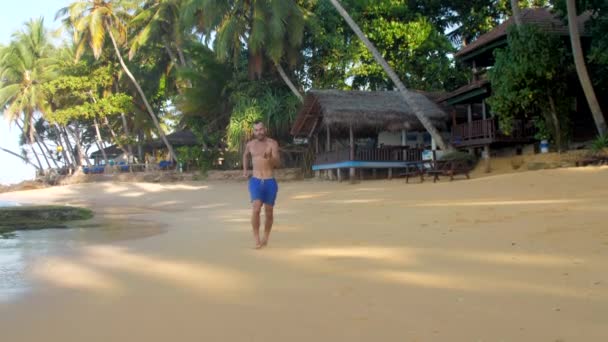 Musclé homme fonctionne sur sable jaune près de bâtiment en bois — Video