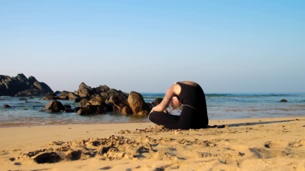 Young woman practices yoga on beach against brown rocks — Stock Video