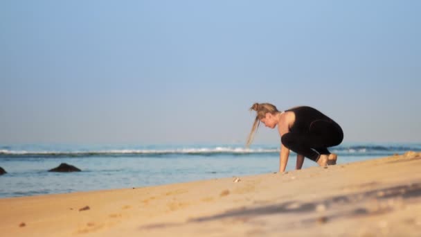 Blondes Mädchen im schwarzen Trainingsanzug übt Yoga am Strand — Stockvideo