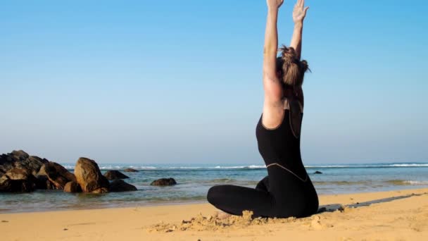 Жінка сидить на піщаному пляжі в позі йоги з піднятими руками — стокове відео