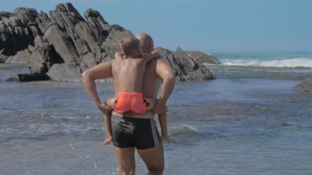 Glücklicher Papa spaziert mit Junge auf dem Rücken am leeren Ozeanstrand entlang — Stockvideo