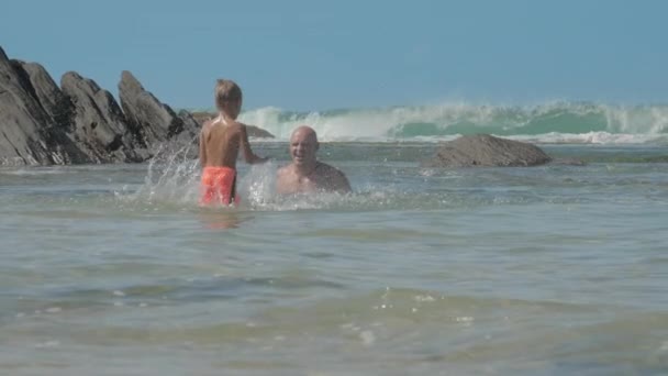 Смішна дитина в шортах бризкає воду на тата в спокійній океанічній бухті — стокове відео
