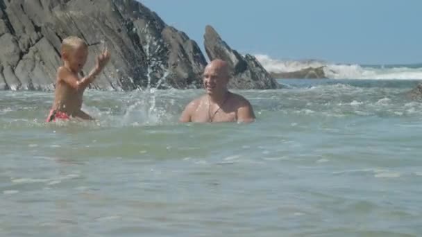 Alegre hombre salpica agua de mar jugando con hijo en rocas — Vídeo de stock