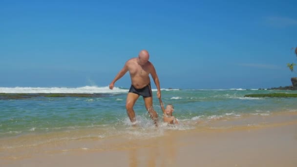 Отец играет с сыном дергая ребенка за руку в океанской воде — стоковое видео