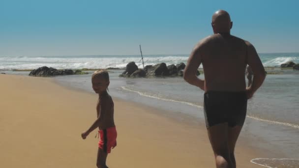 Мальчику с отцом весело бегать по океанскому пляжу к скалам — стоковое видео