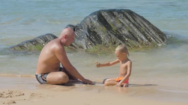 Daddy and cute boy sit on surf line playing with wet sand — Stock Video