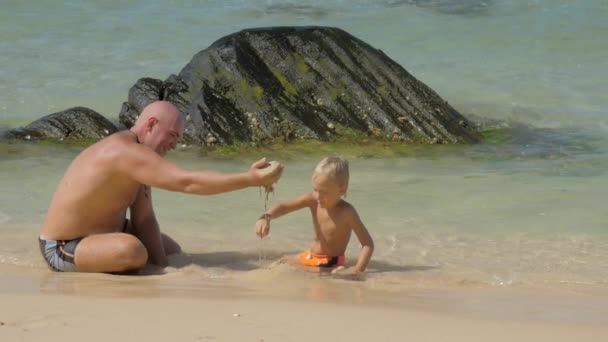 Vader toont nat zand aan grappige zoon rusten in zeewater — Stockvideo