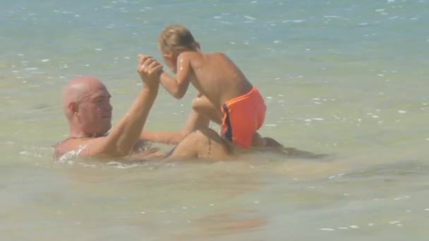 Engraçado menino sobe em pai joelhos jogando em claro oceano baía — Vídeo de Stock