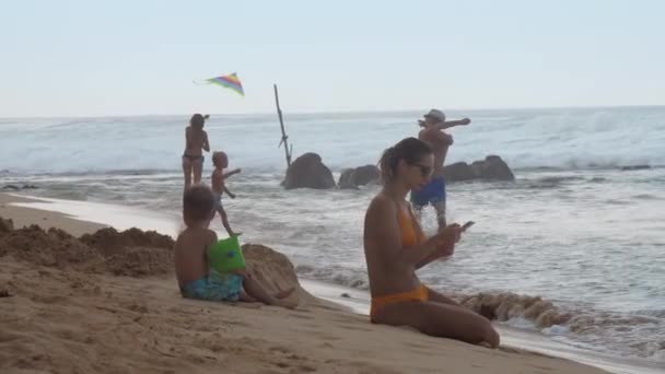 Lady sits on beach sand near boy in arm floats against ocean — стоковое видео