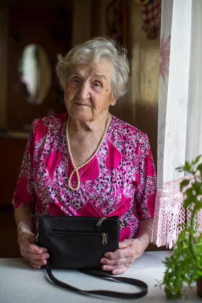 Portrait of an elderly lady with a handbag standing at the table.